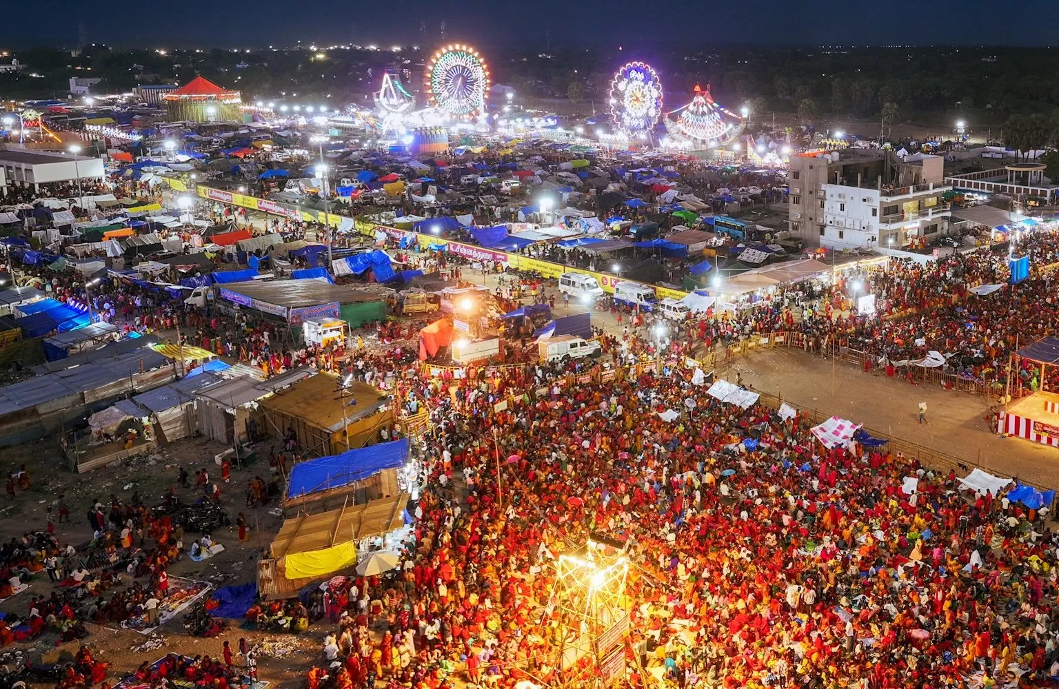Aerial dusk view of a vast festival crowd gathered beside a brightly lit fairground with giant wheels