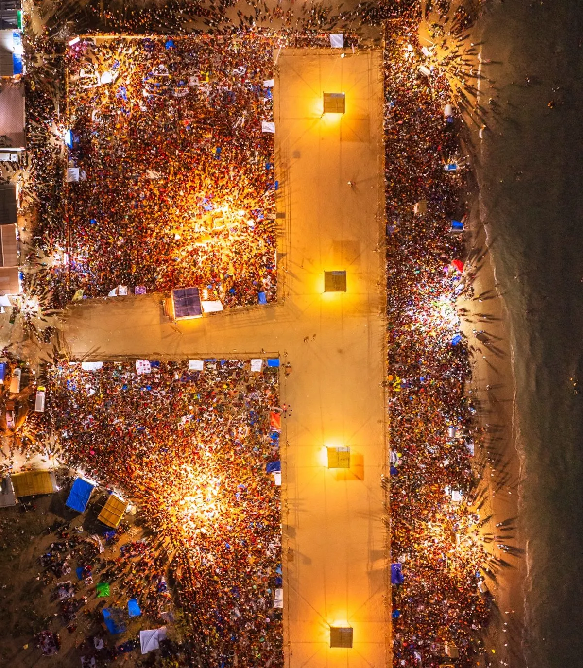 Night drone view looking straight down on dense crowds packed along a lit pathway beside the sea